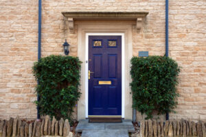 A navy blue front door with brass fixtures, flanked by two green shrubs, set in a beige stone wall of a house.